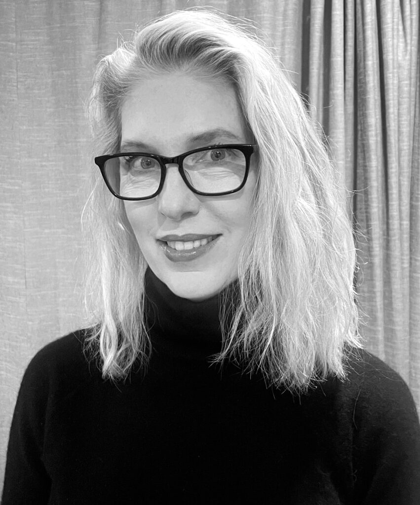 Professional headshot of Melissa Peterson, a woman with shoulder-length wavy light blonde hair, wearing bold dark-framed glasses and a black turtleneck, smiling confidently at the camera against a neutral curtain backdrop, in black and white