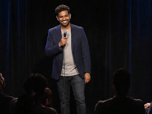 A smiling man with short curly hair and a beard, wearing a dark blazer over a light gray t-shirt and jeans, stands on a dimly lit stage holding a microphone in his right hand. He appears to be performing stand-up comedy, with dark curtains as a backdrop and the silhouettes of audience members visible in the foreground.